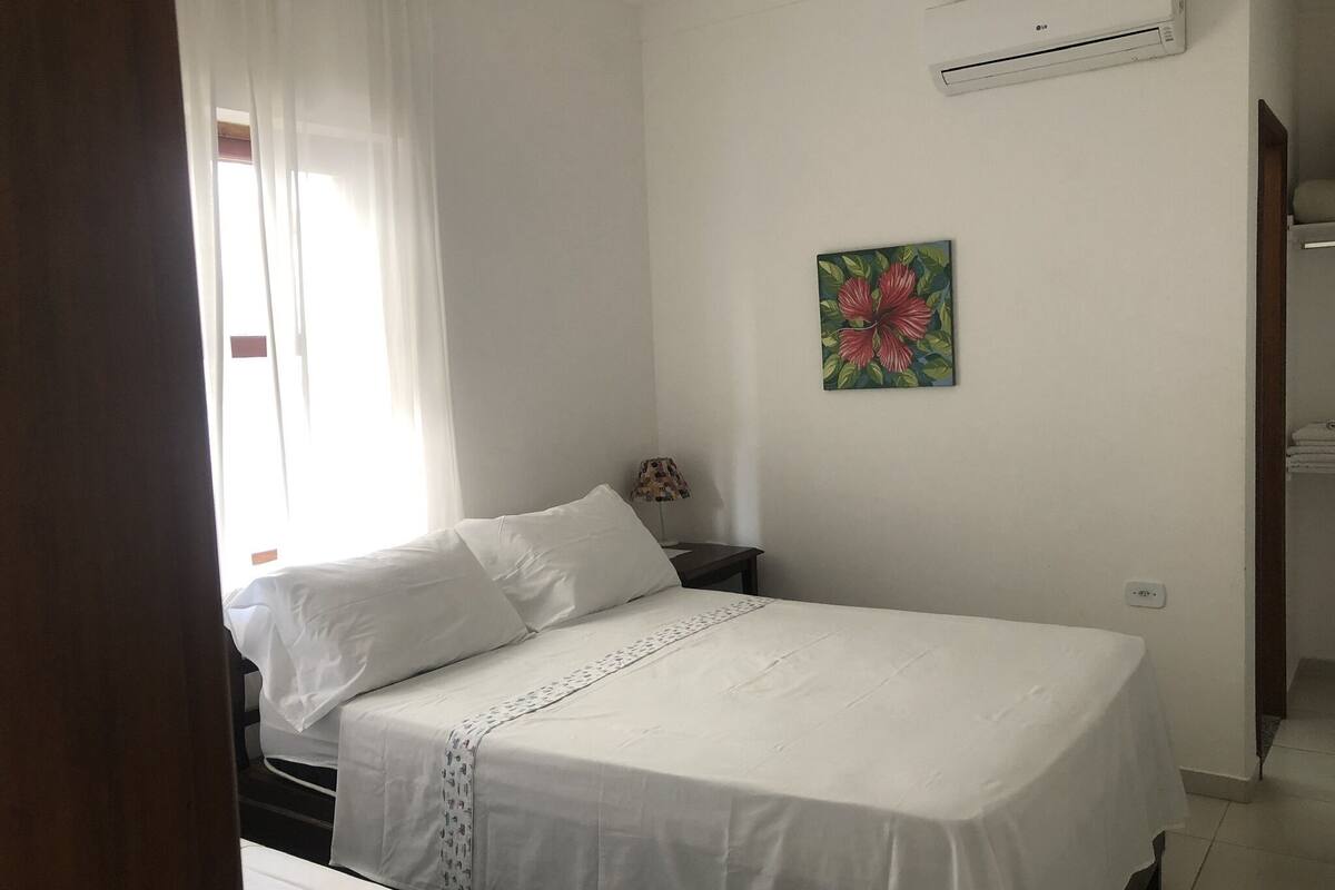 A bedroom with a bed, an air conditioning unit, a nightstand, a painting, and a door leading to another room.