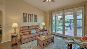 A spacious living room with a brown leather sofa, a wooden coffee table, and a beige armchair. There are two lamps on side tables, a ceiling fan, and a large sliding glass door leading to an outdoor patio. - Tucson Getaway w/ Yard, Pool, Hot Tub + Gas Grill! (Tucson)