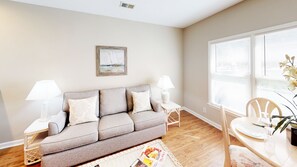 A living room with a grey sofa, two side tables with lamps, a framed picture on the wall, and a dining area with a chair and table.
