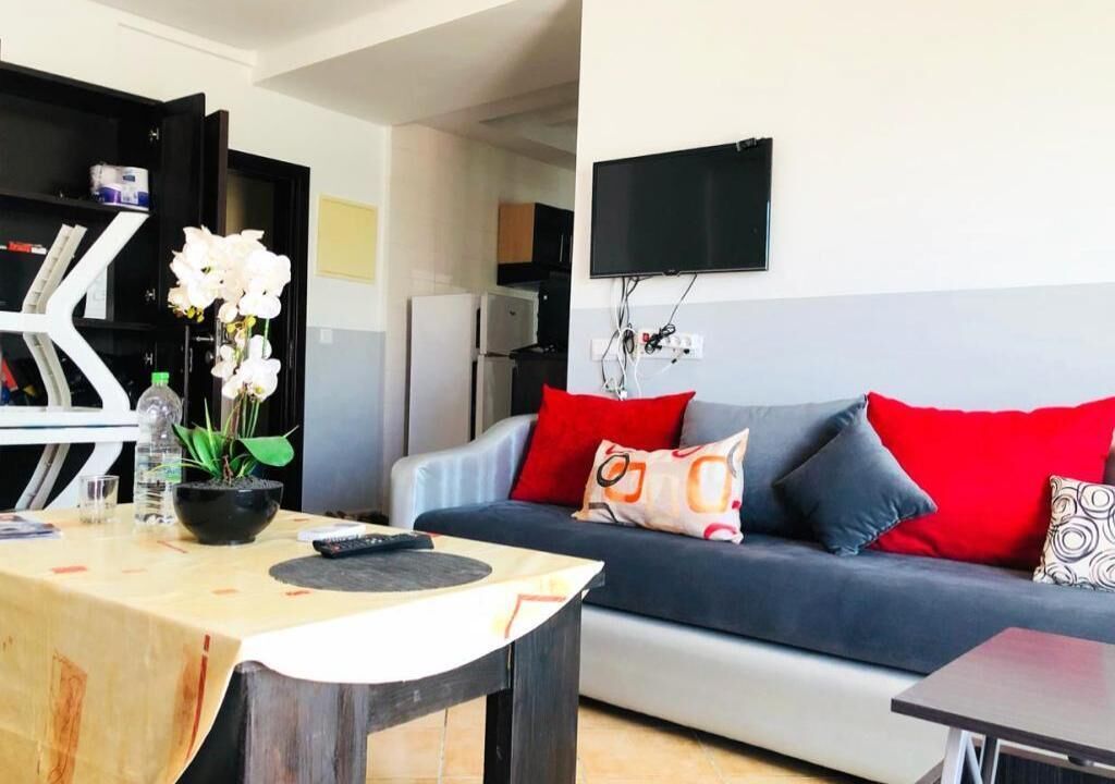 A living room with a sofa, a TV mounted on the wall, and a small table with a remote control.