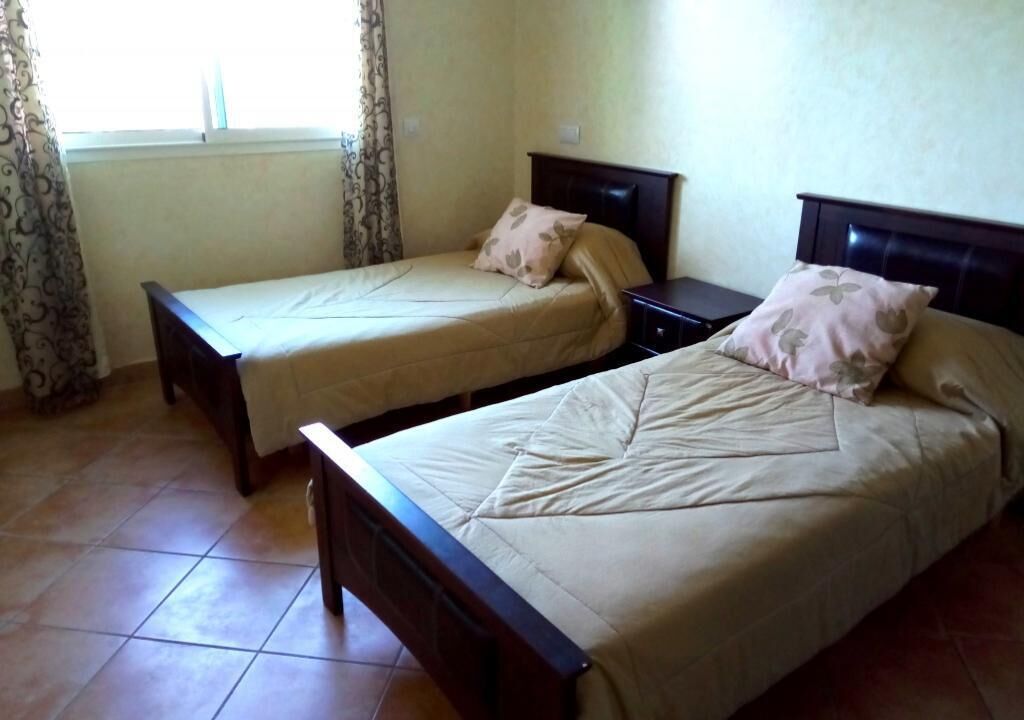 A room with two single beds, each with a wooden headboard and a beige bedspread. There is a window with curtains, and the floor is tiled.