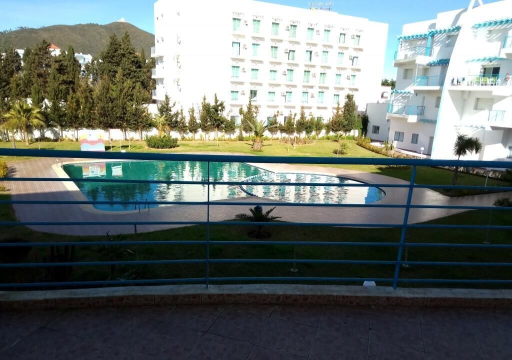 En swimmingpool med et buet design, omgivet af et gelænder. I baggrunden ses flere etagebygninger med altaner og et kuperet landskab.