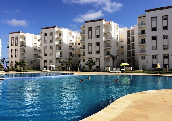 Une piscine entourée d’immeubles à plusieurs étages dotés de balcons, de palmiers et de chaises longues.