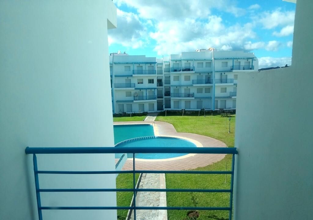 A view of a swimming pool with a curved edge, surrounded by a grassy area and a paved walkway. In the background, there are multi-story buildings with balconies.