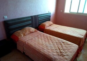 Une chambre avec deux lits simples, une tête de lit sombre et une fenêtre avec des stores.