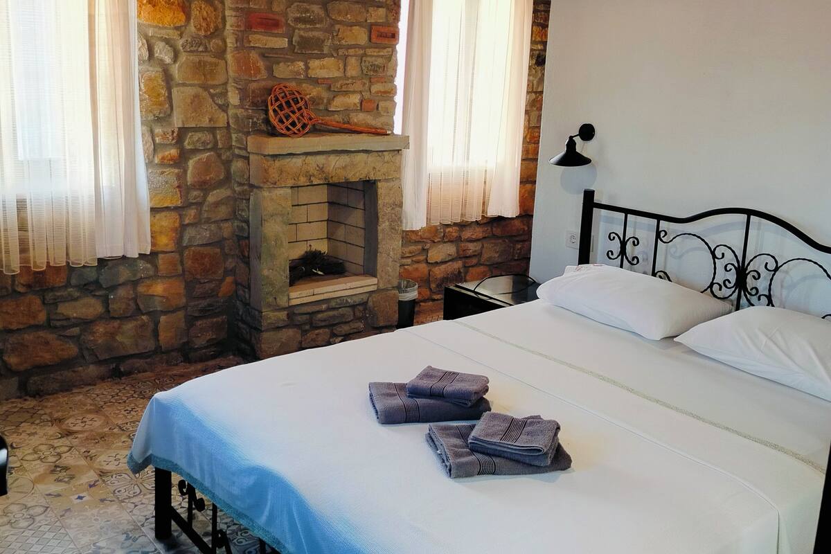 A bedroom with a stone fireplace, a bed with white linens, and a nightstand with a lamp.