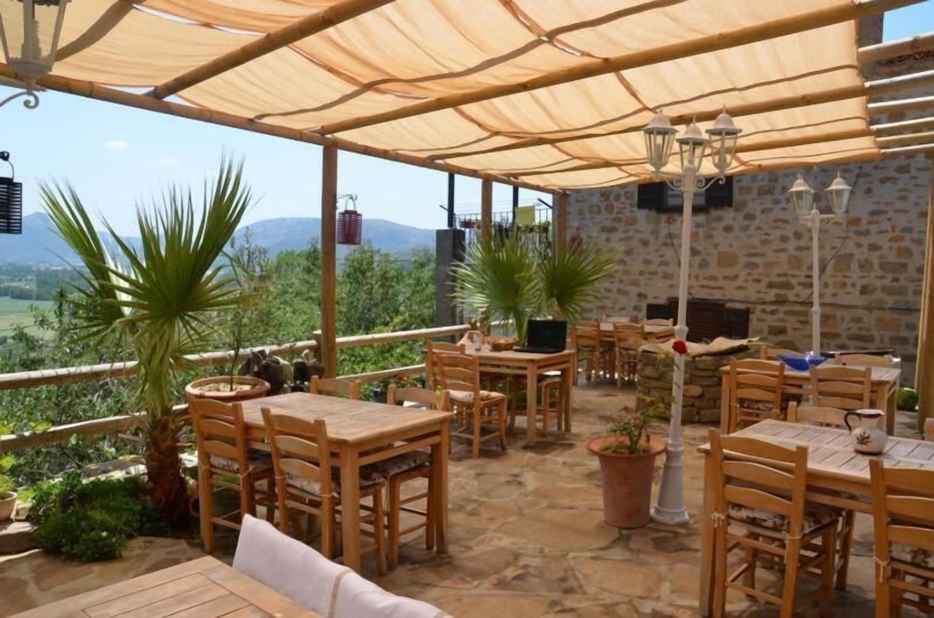 An outdoor patio with wooden tables and chairs, stone flooring, potted plants, and a stone wall backdrop.