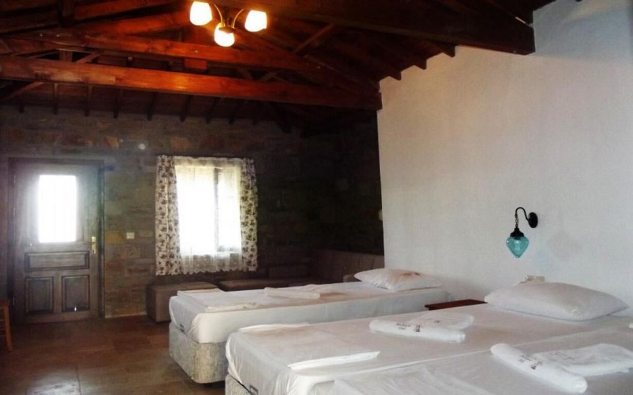 A rustic room with two single beds, a stone wall, a wooden ceiling, and a window with curtains.