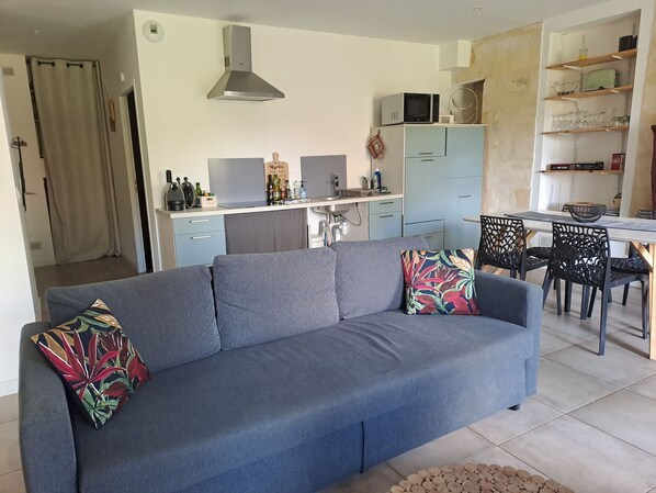 Ein Wohnzimmer mit grauem Sofa, einem Esstisch mit Stühlen, einer Küchenzeile inklusive Schränke und Geräte sowie einem Deckenventilator.
