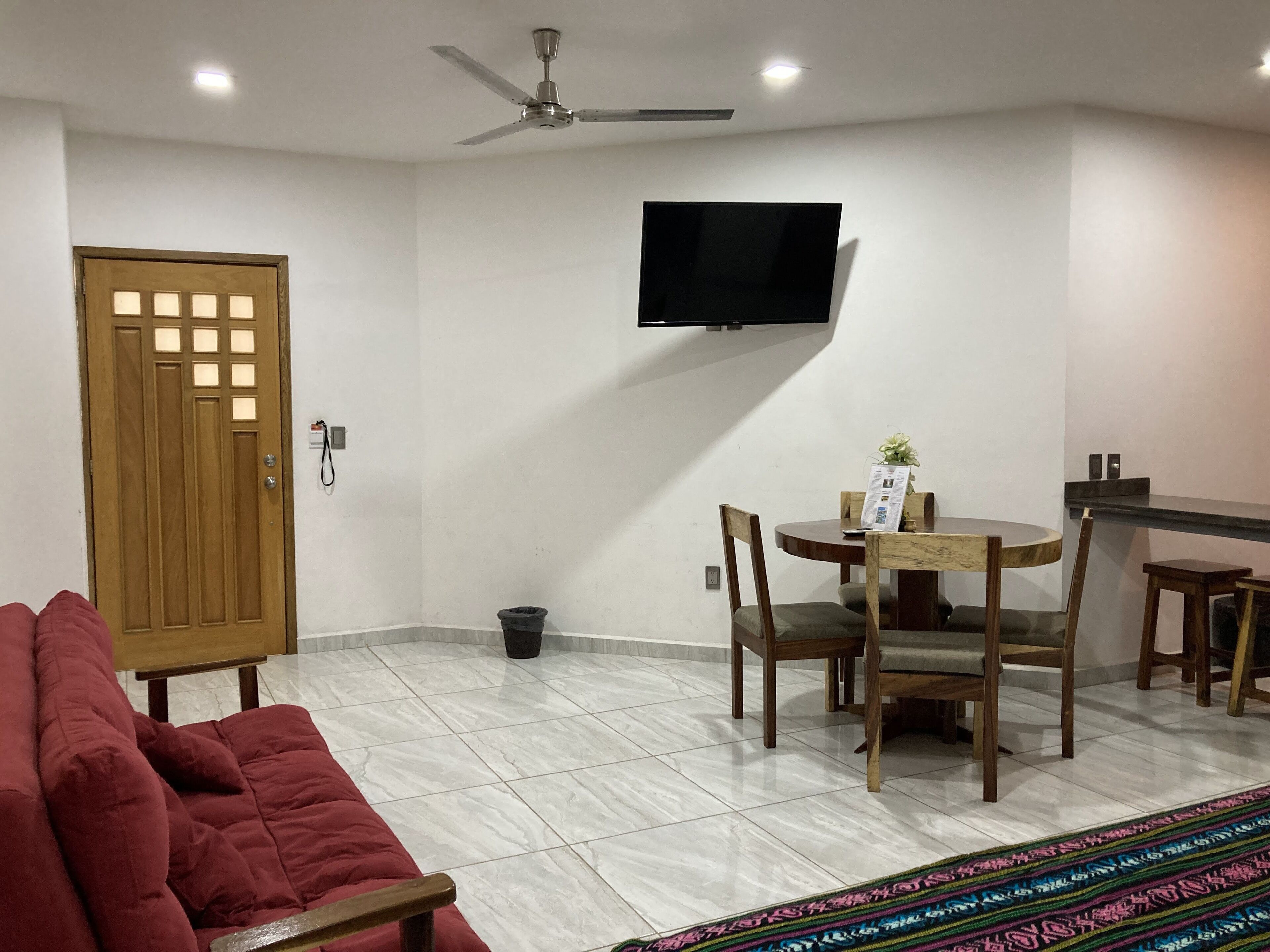 Una habitación de hotel con un sofá rojo, una mesa de comedor con sillas, un ventilador de techo, un televisor montado en la pared y un suelo de baldosas.