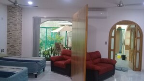 A modern living room with a red sofa, a blue sofa, and a ceiling fan. There is a large window with a view of greenery outside. - PALMAS DE CHAHUE 1 (700 SF) (Bahías de HUATULCO)
