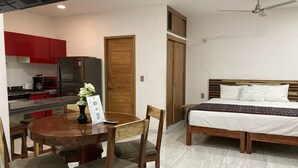 A compact hotel room with a bed, a wooden headboard, a dining table with chairs, a small kitchen area with red cabinets, and a ceiling fan. - PALMAS DE CHAHUE 1 (700 SF) (Bahías de HUATULCO)