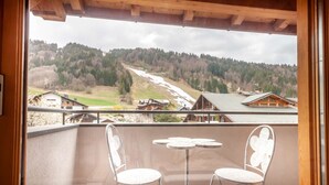 A balcony with two white metal chairs and a small round table, overlooking a mountainous landscape with houses and a ski slope.