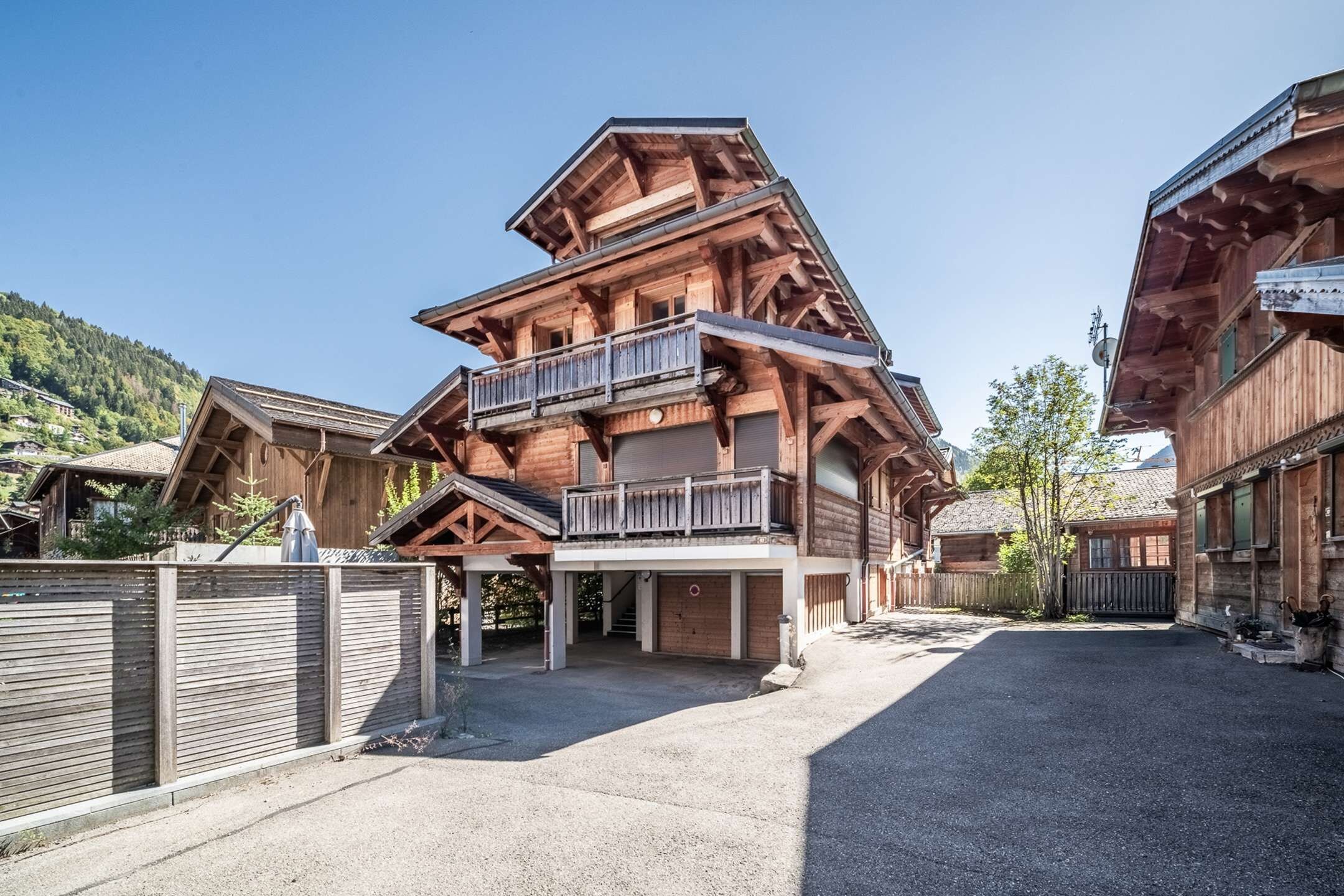 A multi-story wooden building with balconies, a covered parking area, and a mountainous backdrop.