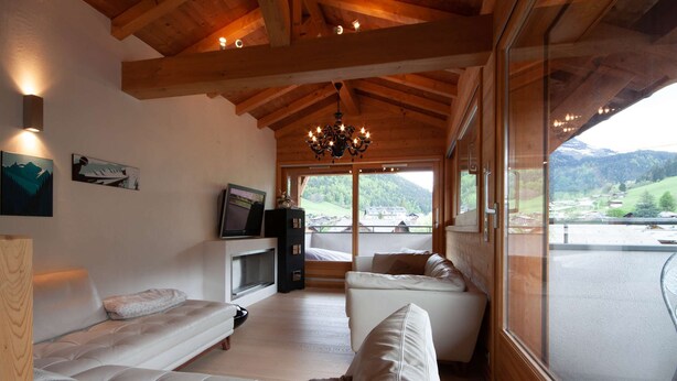 A spacious living room with a large window offering a view of a mountainous landscape. There are two white sofas and a television on a wooden stand.