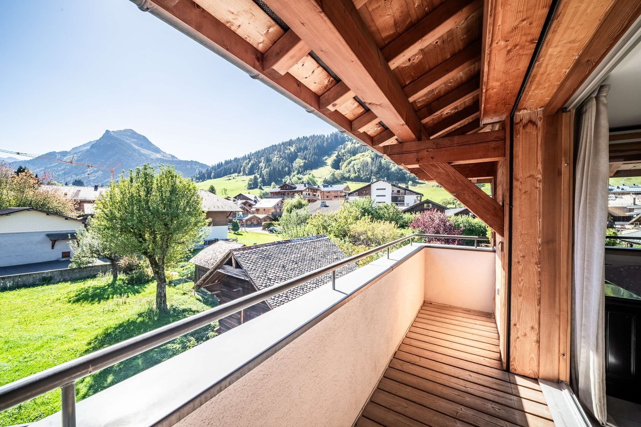 A balcony with a wooden floor, a metal railing, and a view of a village with houses and mountains in the background.