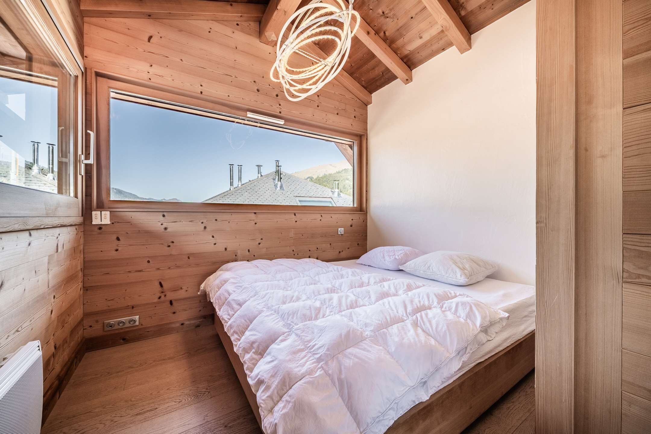 A bedroom with a large window, wooden walls, a bed with white bedding, and a ceiling light fixture.