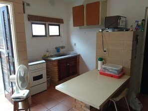 A compact kitchen with a stove, sink, and a small countertop. There is a fan on a stool, a window with blinds, and a shelf with cleaning supplies.