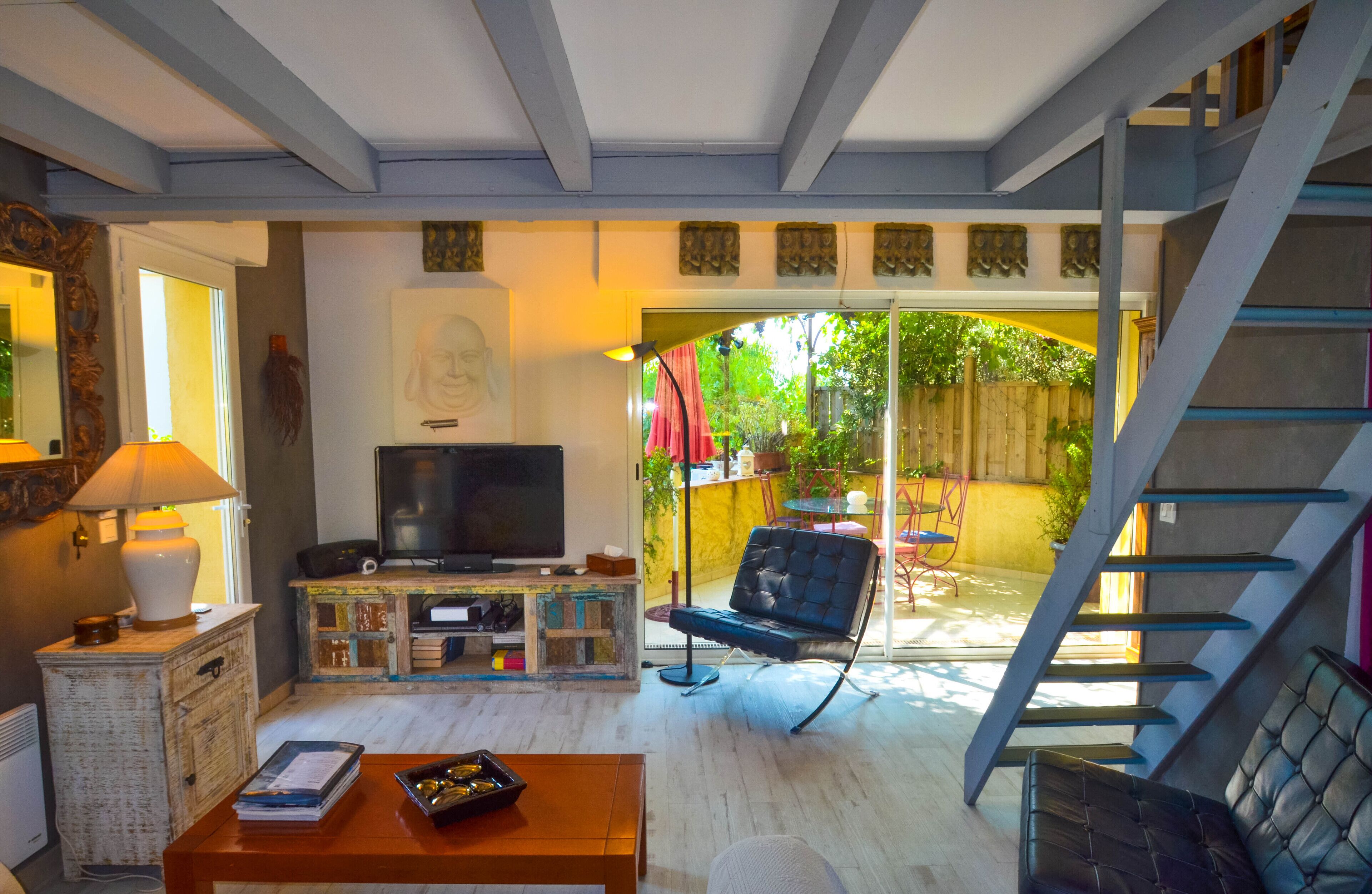 Un salon comprenant un téléviseur, un canapé et une table basse. Un escalier mène à un espace extérieur offrant une vue sur la verdure.