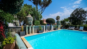 A swimming pool with a stone wall and decorative statues, surrounded by lush greenery and trees.