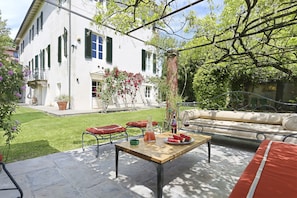 A patio area with a table set for dining, a sofa, and a white building with green shutters in the background.