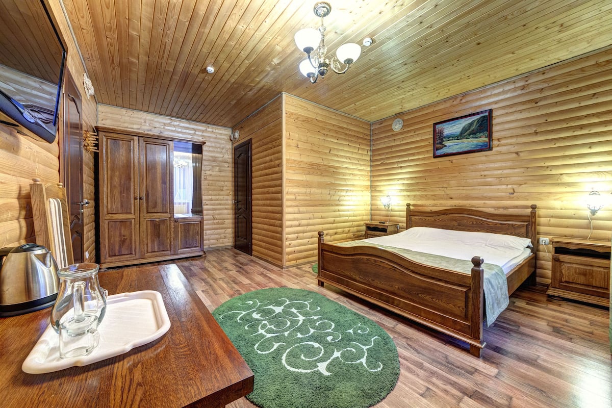 A wooden-paneled room with a bed, a wooden table with a pitcher, and a green rug.