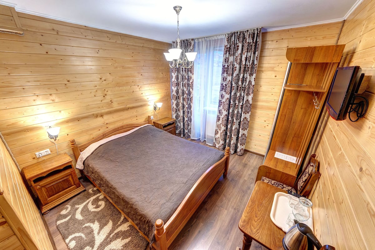 A wooden-paneled room with a bed, bedside table, lamp, and a TV mounted on the wall.