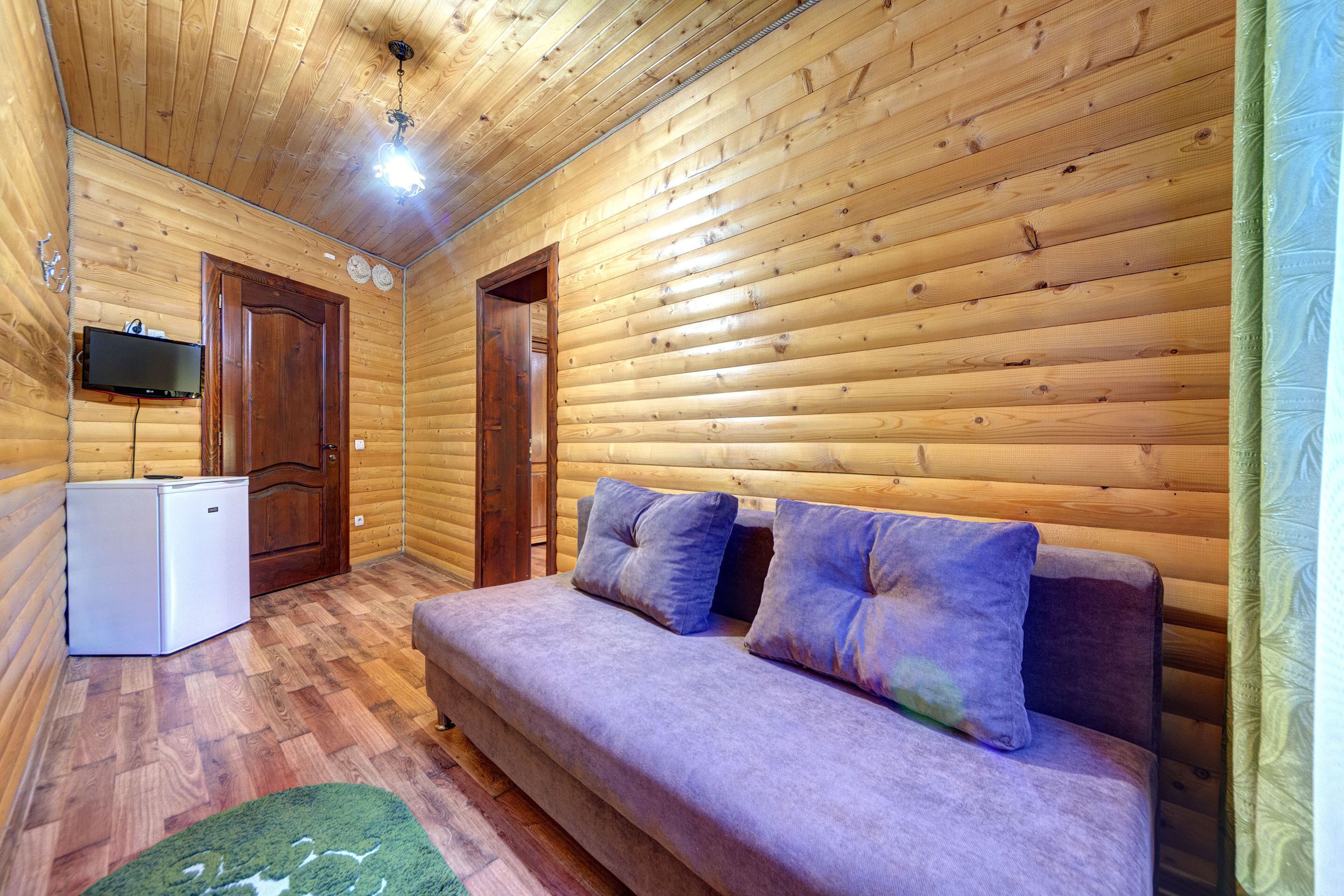 Interieur van een blokhut met een houten plafond, een kleine witte koelkast, een paarse bank met kussens en een wandgemonteerde televisie.