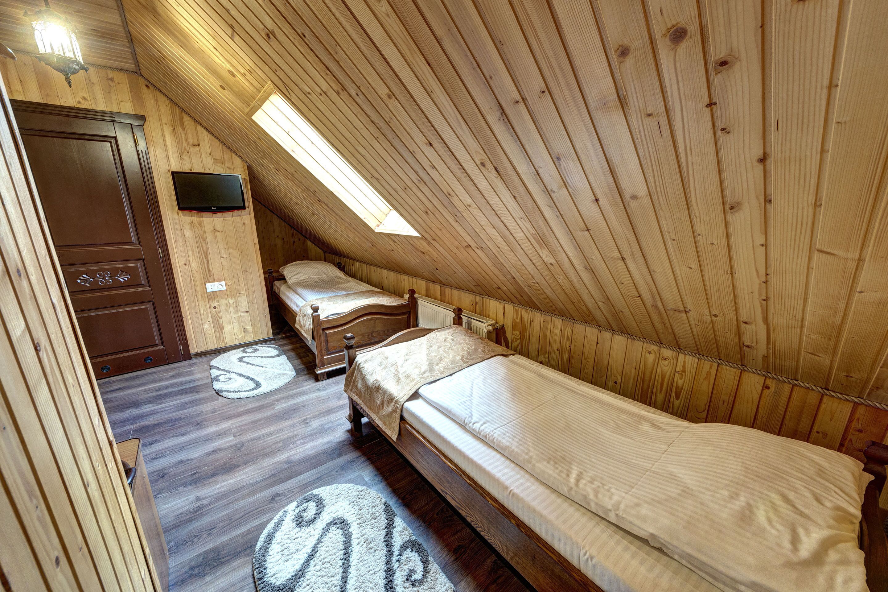 A cozy attic bedroom with wooden walls, a sloped ceiling, a large window, a bed, a dresser, and a television.