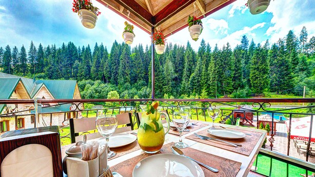 A dining table set with plates, cutlery, and a pitcher of iced tea, overlooking a forested landscape. - Hotel Milli & Jon (Bukovel)