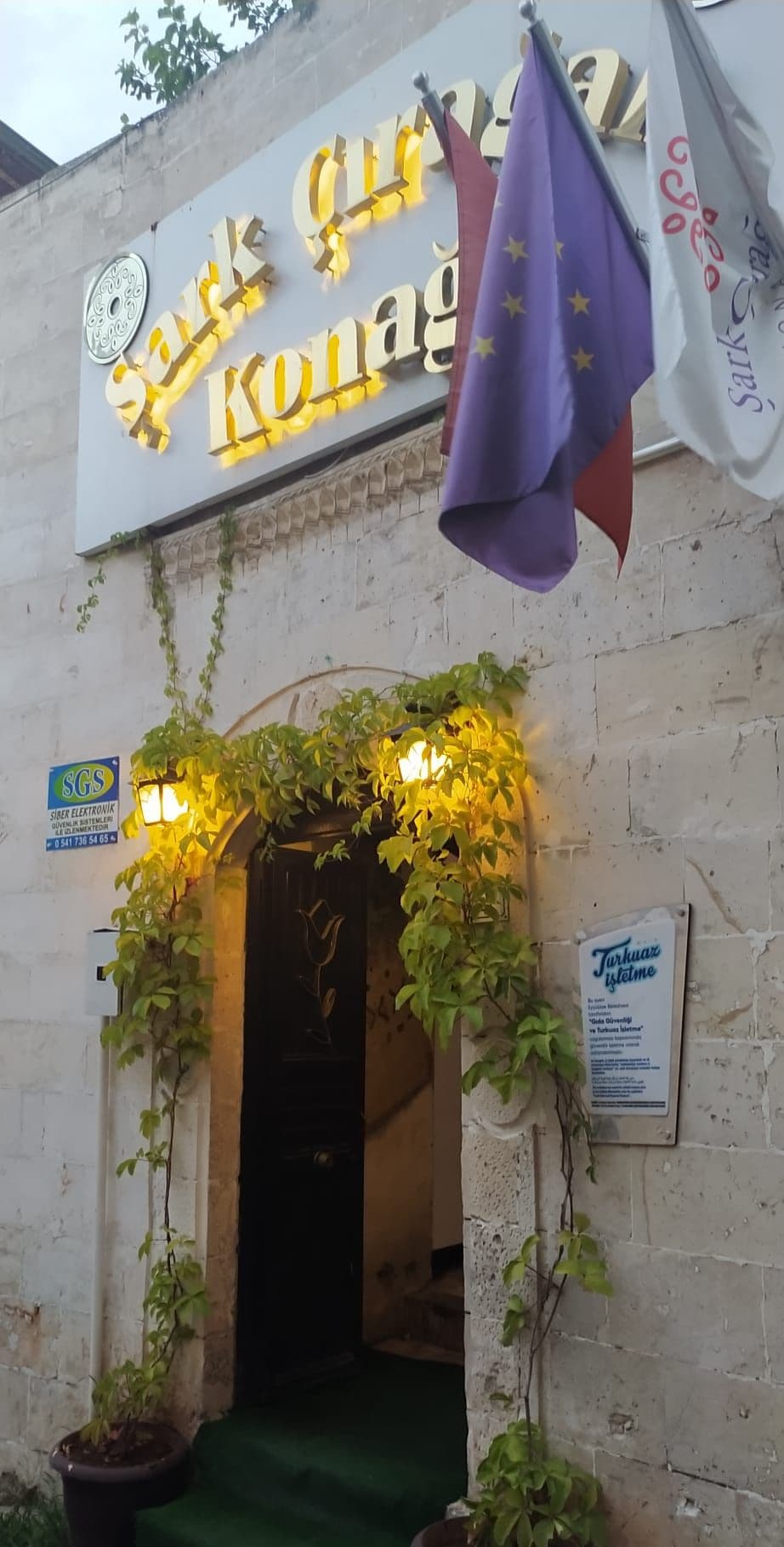 Entrada de un edificio con un letrero que dice 'Saray Konak' y una bandera. Hay plantas y un letrero de 'SGS' en la pared.