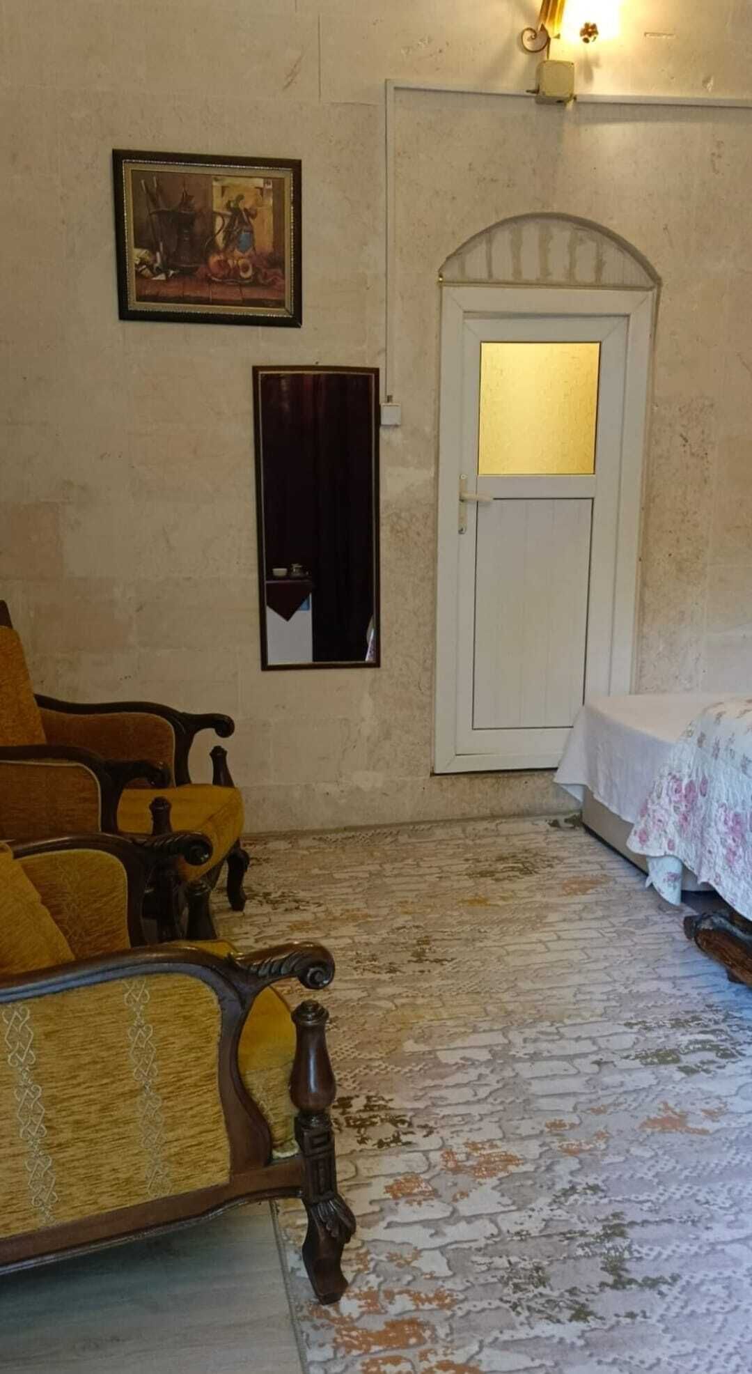 Una habitación con una silla de madera, una cama con cubrecama floral, una puerta con un pequeño ventanal y un cuadro en la pared.