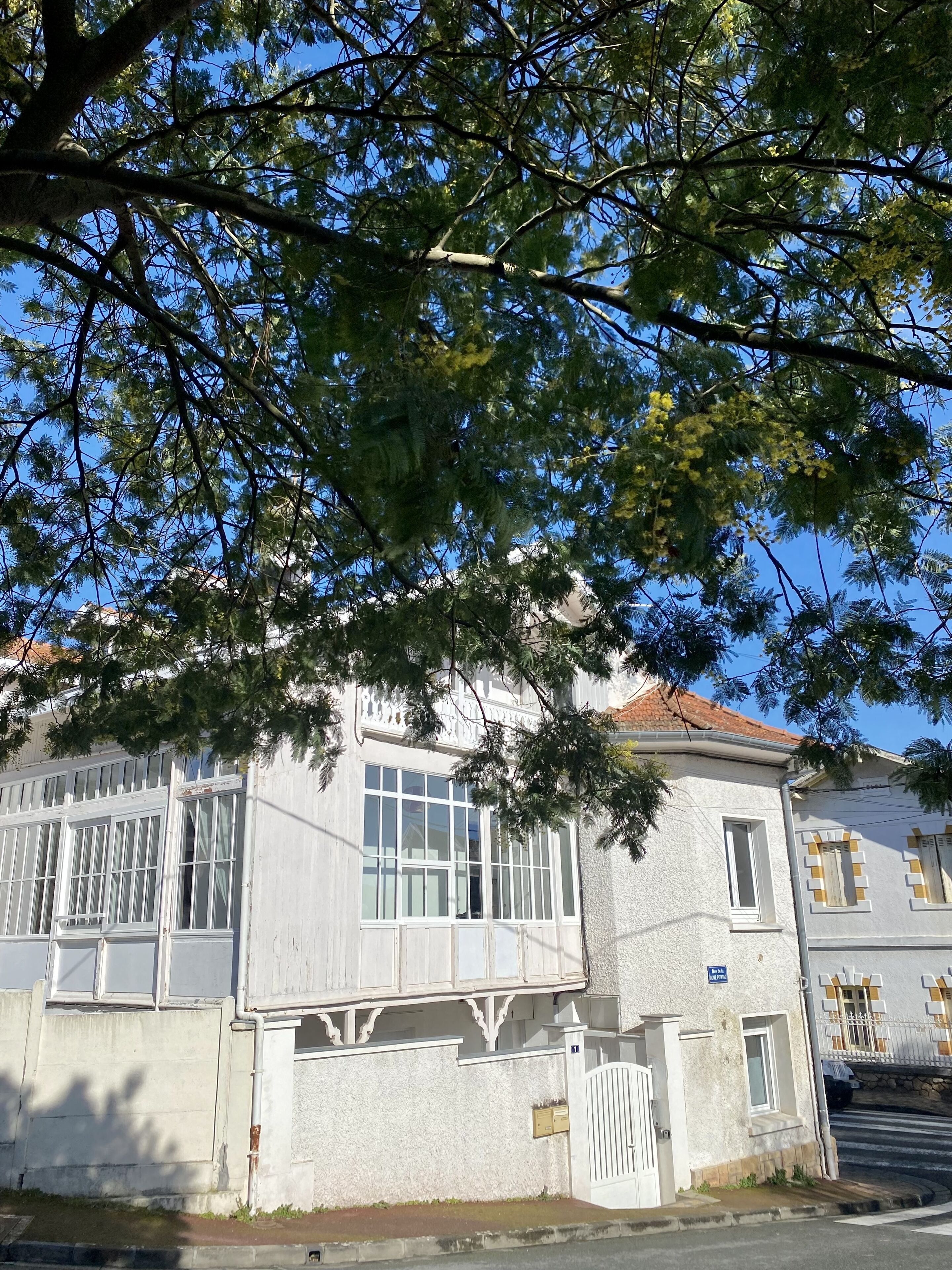 A white building with large windows and a balcony, surrounded by trees.
