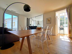 A modern dining area with a wooden table and clear chairs, a kitchen in the background, and large windows allowing natural light.