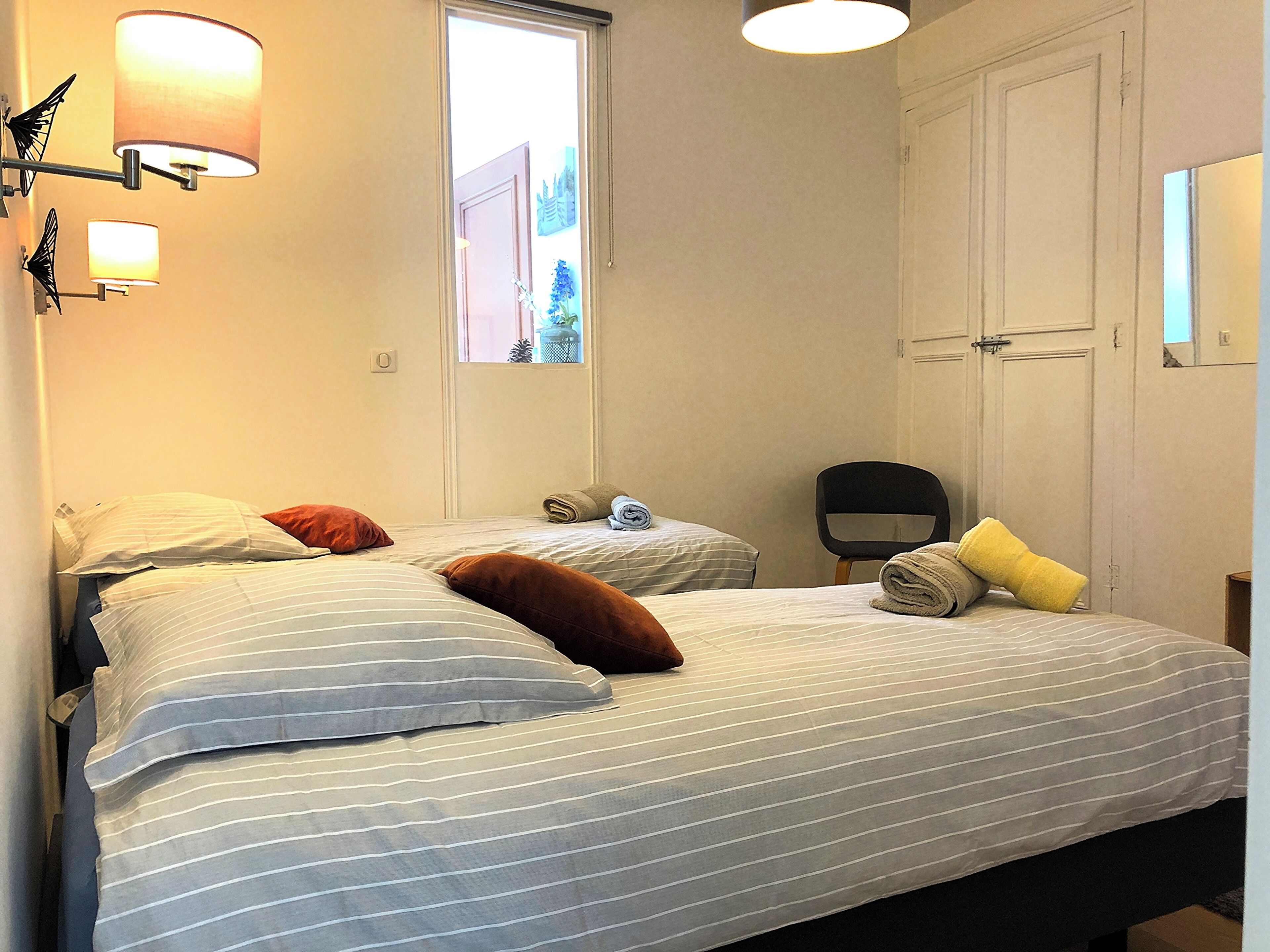A bedroom with two beds, a window, a chair, and wall-mounted lamps.