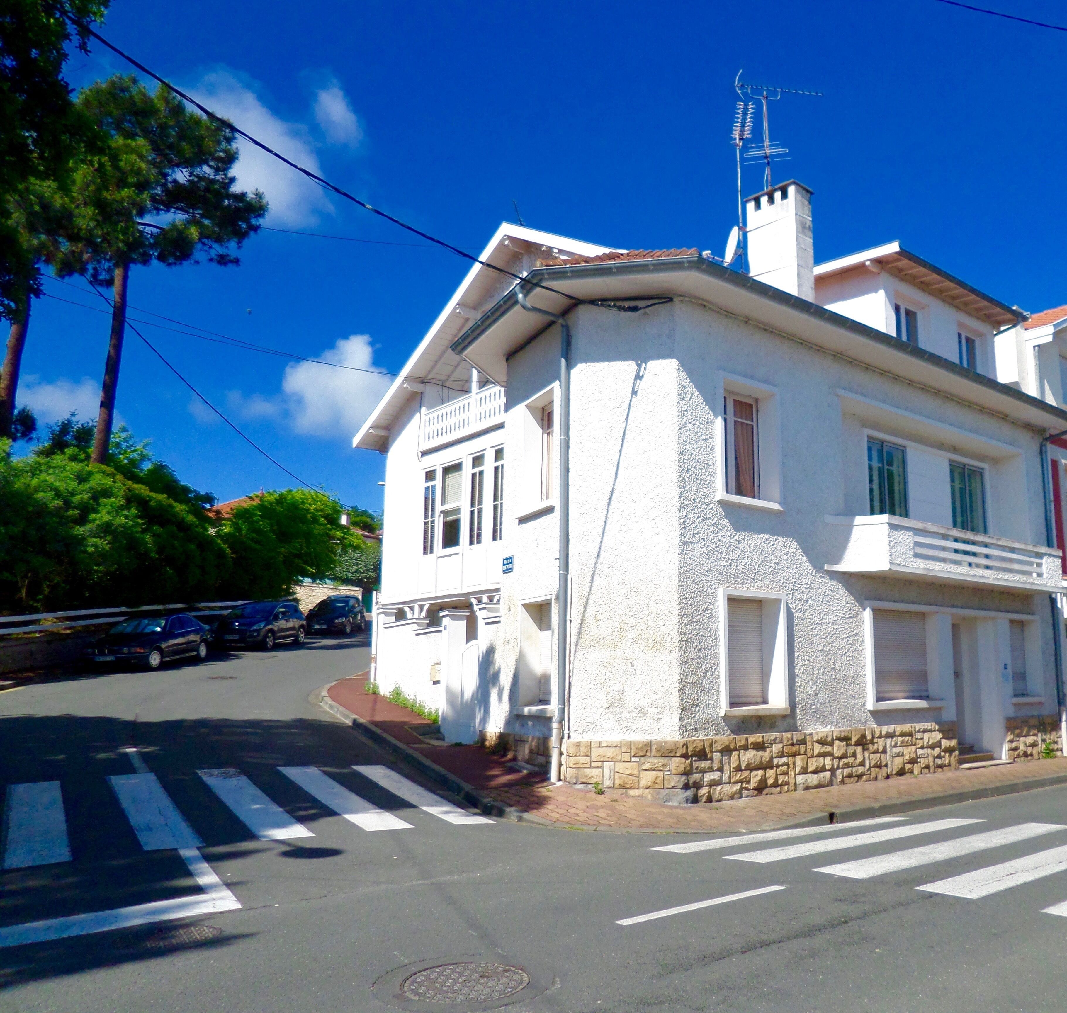 A white building with a stone base, a balcony, and a street view with cars and a crosswalk.