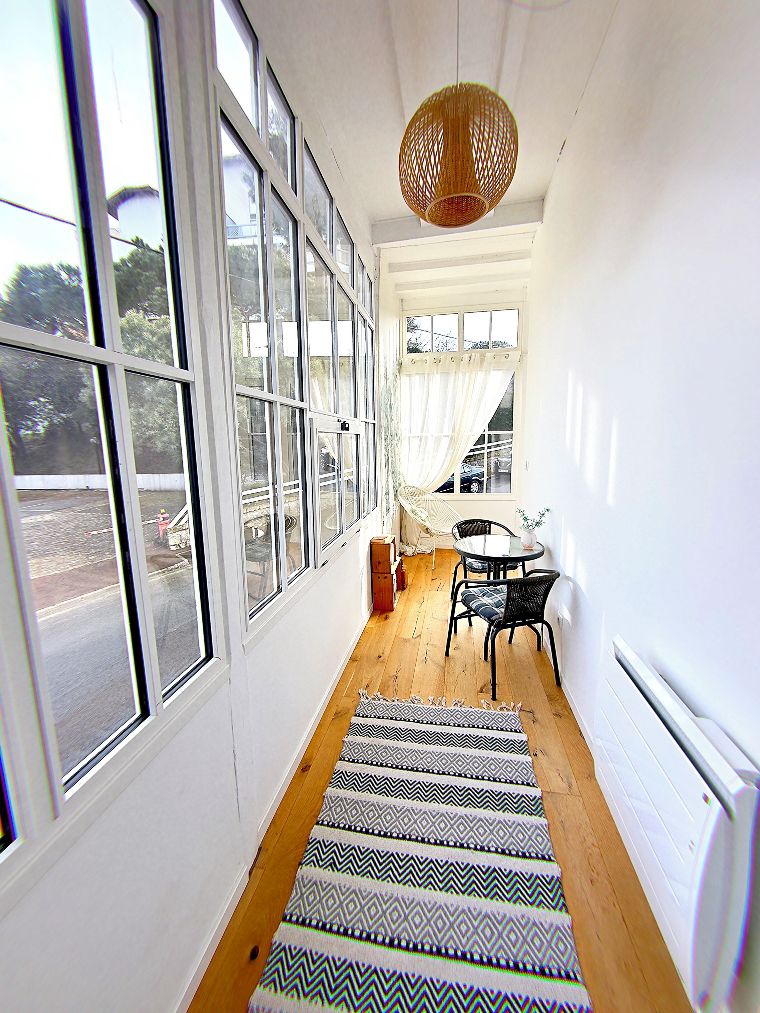 A narrow hallway with large windows, a dining area with a table and chairs, and a wicker hanging light fixture.