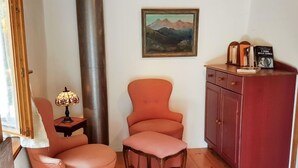 A room with a wooden floor, a red cabinet, a pink armchair, a smaller pink chair, a table with a lamp, and a framed painting on the wall.