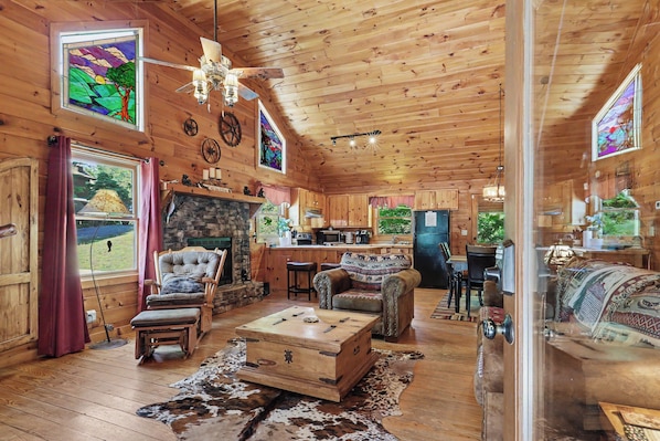 A wooden cabin interior with a stone fireplace, leather furniture, and a kitchen area.