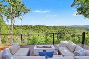An outdoor deck with a fire pit, seating area, and a view of a forested landscape.