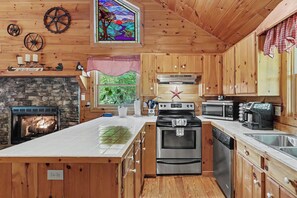 A wooden cabin kitchen with a stone fireplace, wooden cabinets, a large island, and a view of greenery outside. - LAKE BLUE RIDGE VIEW & ACCESS, POOL TBL, PROJECTOR, FIRE PIT, HOT TUB, DOGS OKAY (Blue Ridge)