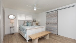 A bedroom with a bed, a bedside table with a lamp, a wooden bench, a ceiling fan, and a sliding barn-style door. - Beachy Getaway–Cozy, Clean & Equipped w/ All Essentials! Relax, Enjoy & Refresh! (St. Augustine)