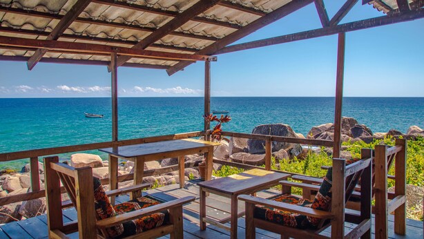 A wooden deck with seating, overlooking a clear blue sea with rocks and a small boat. - Ulisa Bay Lodge (Likoma Island)