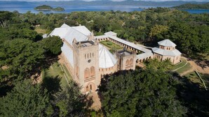 A historic building with a central tower and multiple smaller towers, surrounded by lush greenery and a body of water in the background. - Ulisa Bay Lodge (Likoma Island)