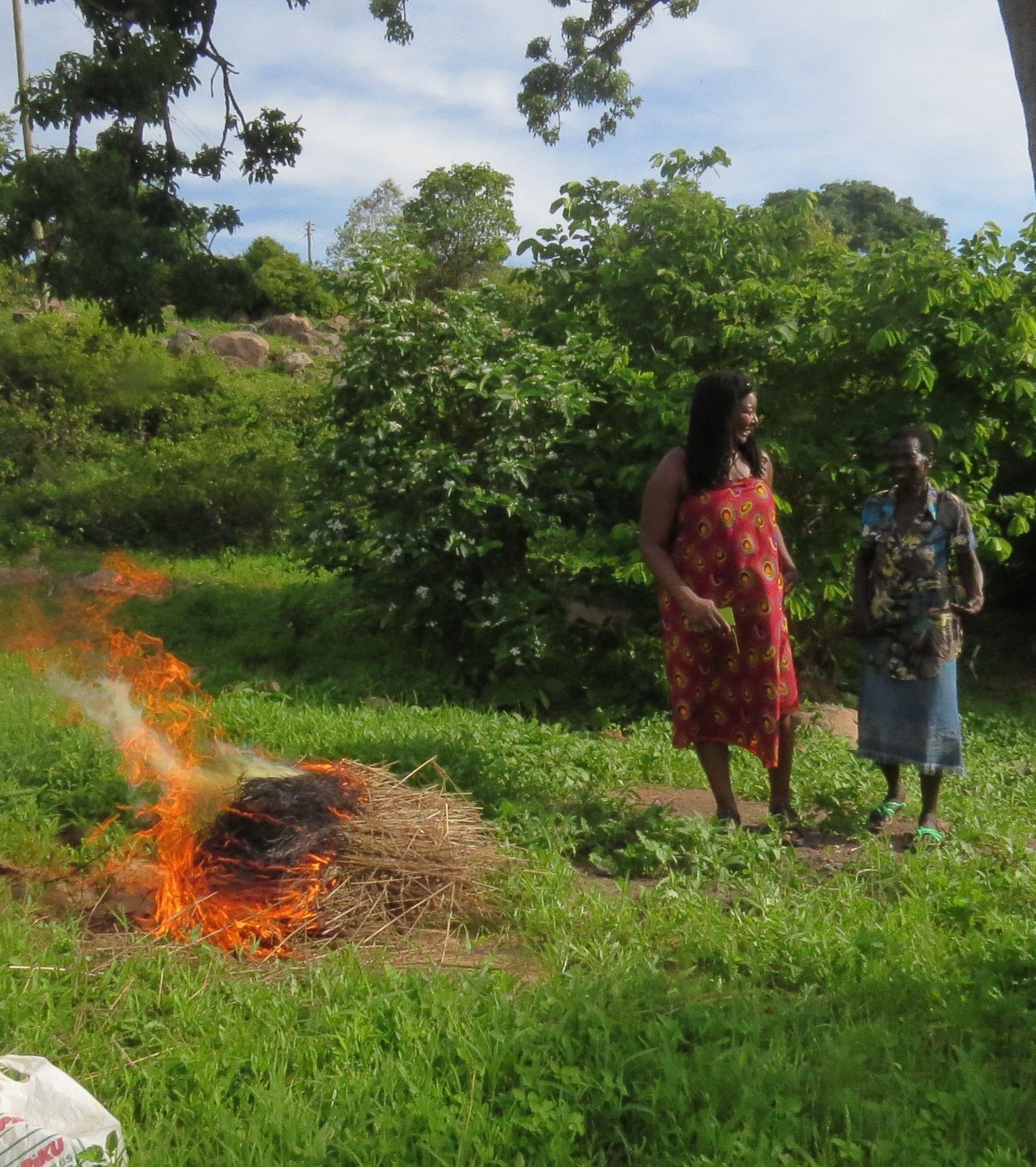 A person in a red dress standing next to a fire in a grassy area with trees in the background.