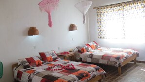 A room with two wooden beds, each with patterned bedding and pillows. There are wall-mounted lights and a window with a floral curtain. - Ulisa Bay Lodge (Likoma Island)
