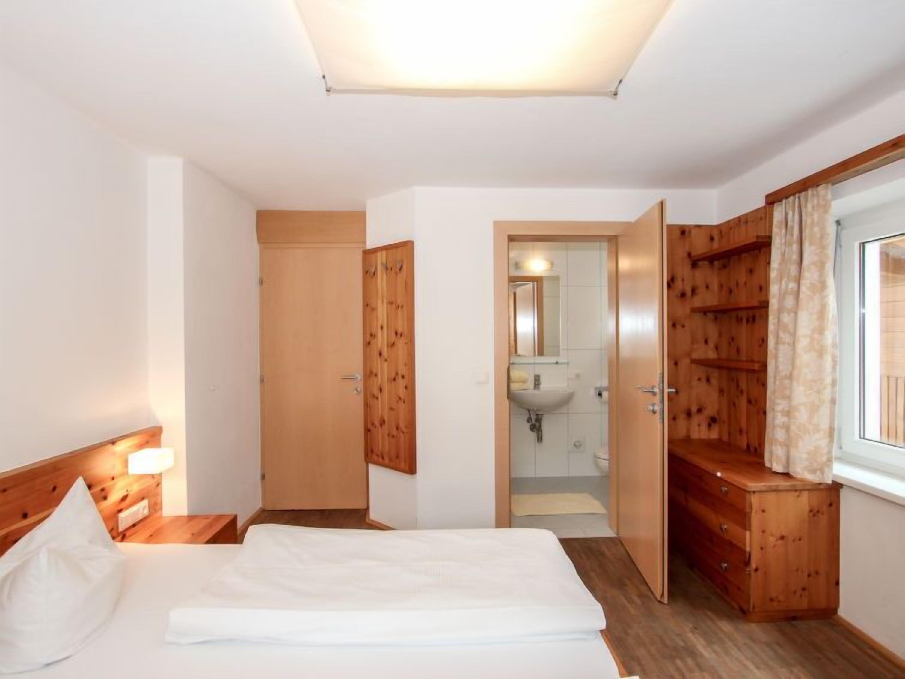 A hotel room with a bed, wooden wardrobe, bedside table, and a bathroom visible through an open door.