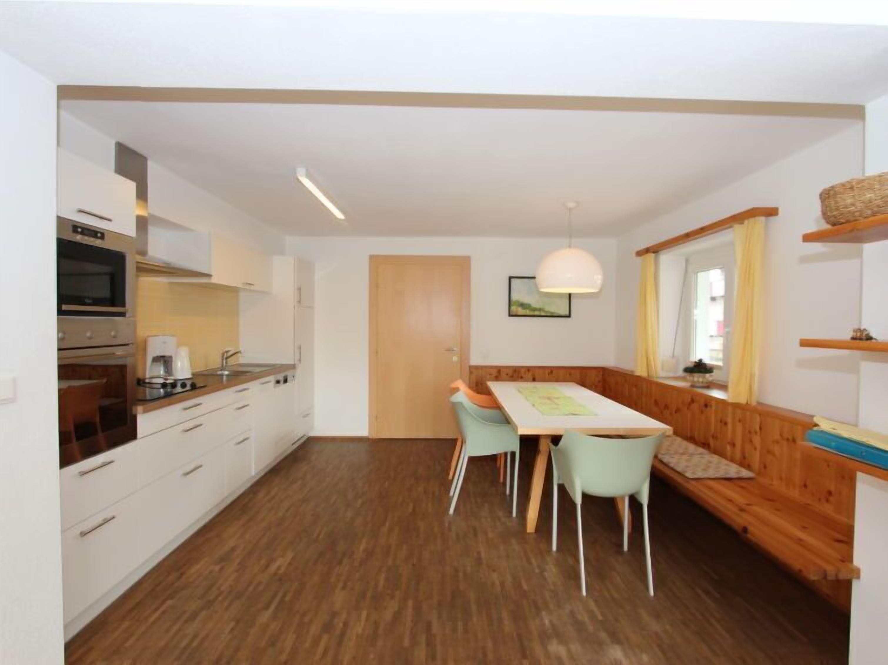 A compact kitchen with white cabinets, a built-in oven, and a dining area with a wooden bench and two chairs.