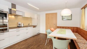 A modern kitchen with white cabinets, a built-in oven, and a coffee maker. There is a wooden door and a bench with cushions in the dining area. - Apartment Gerda by Interhome (Kaltenbach)