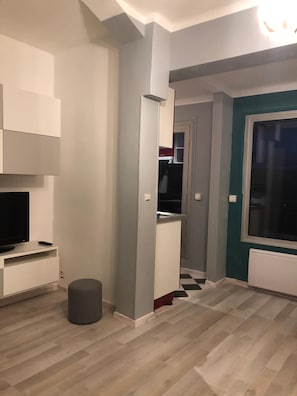 A modern living room with a flat-screen TV, a small ottoman, and a hallway leading to a kitchen area. - Beautiful apartment with balcony and panoramic views of the mountains (Font-Romeu-Odeillo-Via)
